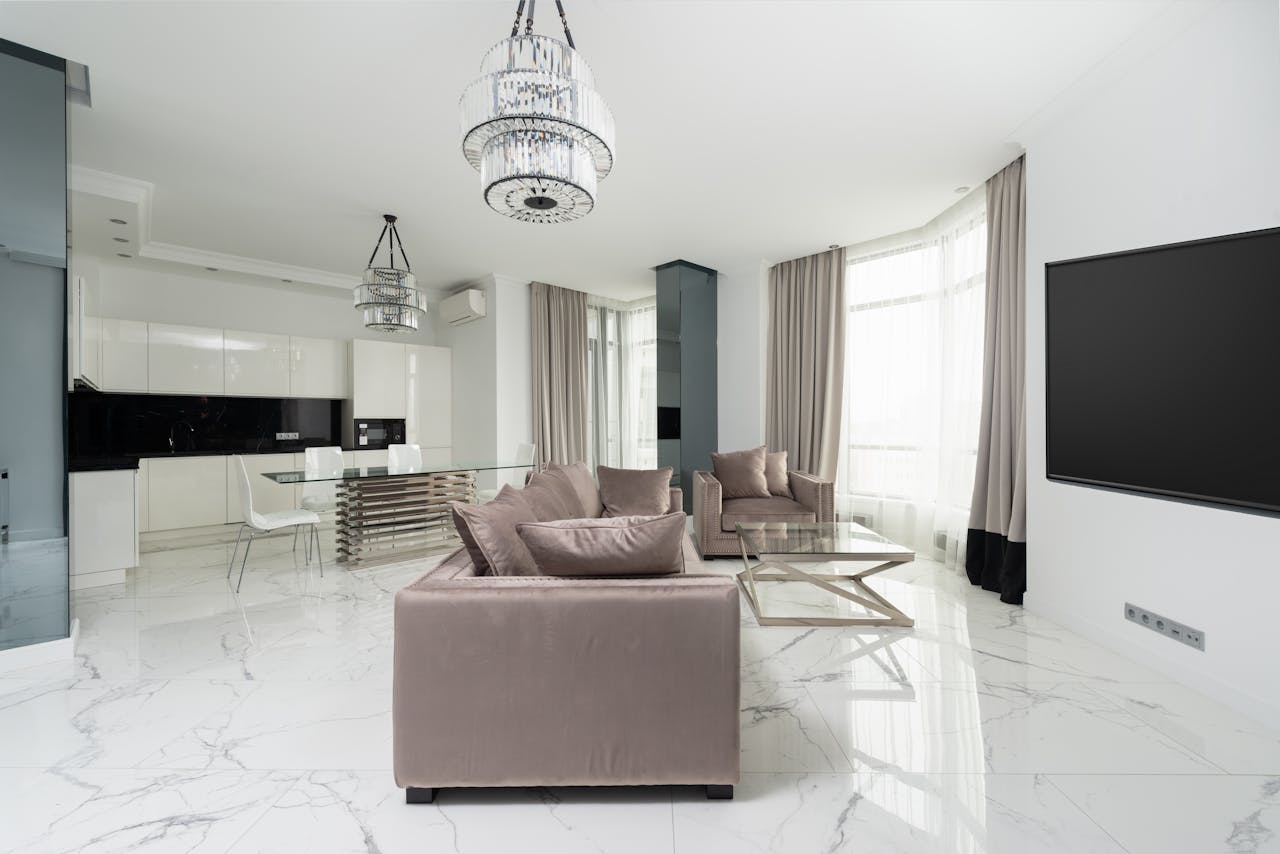 Interior of modern light studio with couch and armchair with pillows near glass table and chairs under chandelier next to open kitchen with white cabinets and windows with curtains